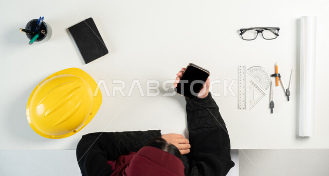 The concept of engineering and architectural construction, obtaining the necessary supplies via the Internet, shopping via websites, a vertical overhead portrait of a Saudi Arabian Gulf woman using a phone, next to her is a protective helmet, engineering tools, plans, and medical glasses, a white background.