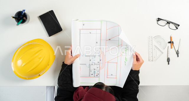 Implementing construction projects, the concept of architectural engineering and construction, obtaining the necessary supplies via the Internet, shopping via websites, a vertical overhead portrait of a Saudi Arabian Gulf woman holding an engineering diagram and next to her a protection helmet and engineering tools, white background.