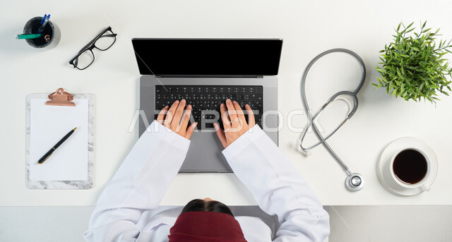 التسوق عبر المواقع الالكترونية، مفهوم الطب والرعاية الصحية، الخدمات والأجهزة الطبية، التسويق للأدوات الحديثة، بورتريه علوي عامودي لإمرأة عربية خليجية سعودية ترتدي المعطف الطبي تستخدم الحاسوب المحمول، دمج التكنولوجيا بالحياة اليومية، خلفية بيضاء