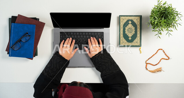 البيع والشراء عبر الانترنت، دمج التكنولوجيا بالحياة اليومية، بورتريه علوي عامودي ليدي إمرأة عربية خليجية سعودية تقوم باستخدام الحاسوب المحمول بجانبها القرآن الكريم والسبحة الصفراء، شراء الكتب الدينية والمصاحف الشريفة من خلال المواقع الالكترونية، خلفية بيضاء