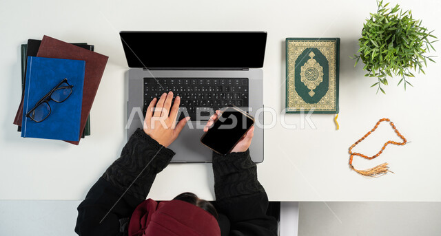 دمج التكنولوجيا بالحياة اليومية، بورتريه علوي عامودي ليدي إمرأة عربية خليجية سعودية تقوم باستخدام الهاتف والحاسوب بجانبها القرآن الكريم والسبحة الصفراء، تصفح المواقع الالكترونية لشراء الكتب الدينية والمصاحف الشريفة، البيع والشراء عبر الانترنت، خلفية بيضاء