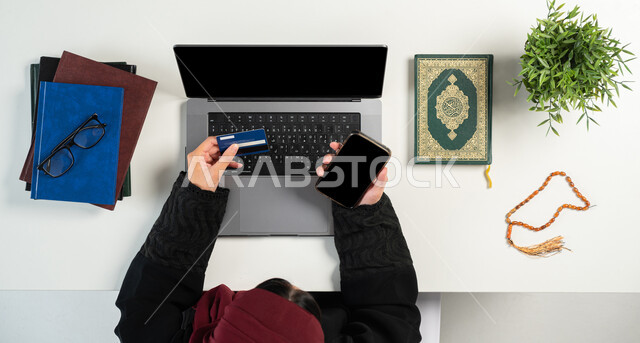 شراء الكتب الدينية والمصاحف الشريفة من خلال المواقع الالكترونية، بورتريه علوي عامودي ليدي إمرأة عربية خليجية سعودية تستخدم الحاسوب والهاتف بجانبها القرآن الكريم والسبحة الصفراء، الدفع عبر البطاقة البنكية، دمج التكنولوجيا بالحياة اليومية، خلفية بيضاء