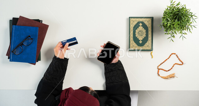 تصفح المواقع الالكترونية لشراء الكتب الدينية والمصاحف الشريفة، بورتريه علوي عامودي ليدي إمرأة عربية خليجية سعودية تستخدم الهاتف المحمول بجانبها القرآن الكريم والسبحة الصفراء، الدفع عبر البطاقة البنكية، دمج التكنولوجيا بالحياة اليومية، خلفية بيضاء