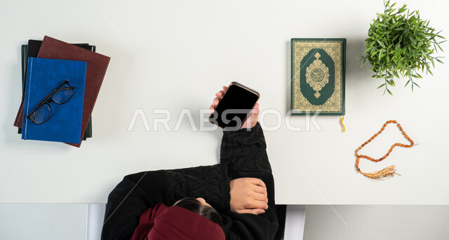 شراء الكتب الدينية والمصاحف الشريفة من خلال المواقع الالكترونية، الطلب والبيع والشراء عبر الانترنت، بورتريه علوي عامودي ليدي إمرأة عربية خليجية سعودية تستخدم الهاتف المحمول بجانبها القرآن الكريم والسبحة الصفراء، دمج التكنولوجيا بالحياة اليومية، خلفية بيضاء