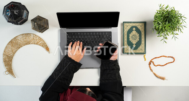 Taking advantage of modern technologies, ordering via websites, a vertical overhead portrait of a Saudi Gulf Arab woman using a computer and phone next to her is the Holy Qur’an, lanterns, and the crescent moon, religious occasions and Islamic holidays, preparing for the advent of the holy month of Ramadan, white background.