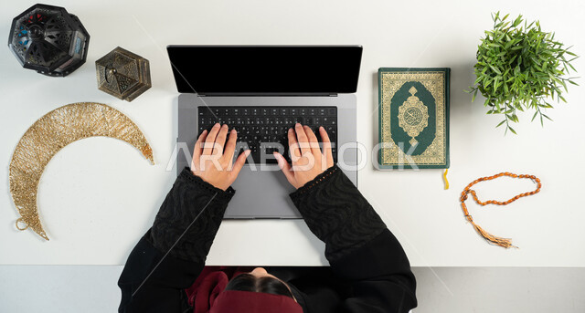 البيع والشراء عبر الانترنت، الإستعداد لقدوم شهر رمضان المبارك، المناسبات الدينية والأعياد الإسلامية، الطلب عبر المواقع الالكترونية، بورتريه علوي عامودي ليدي إمرأة عربية خليجية سعودية تستخدم الحاسوب المحمول بجانبها المصحف الشريف والفوانيس والهلال، خلفية بيضاء