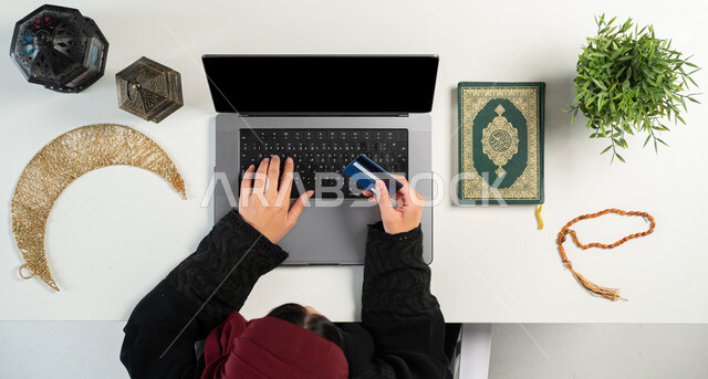 الطلب عبر المواقع الالكترونية، المناسبات الدينية والأعياد الإسلامية، الإستعداد لقدوم شهر رمضان المبارك، بورتريه علوي عامودي ليدي إمرأة عربية خليجية سعودية تستخدم الحاسوب بجانبها المصحف الشريف والفوانيس والهلال، الدفع عن طريق بطاقة الصراف البنكية، خلفية بيضاء