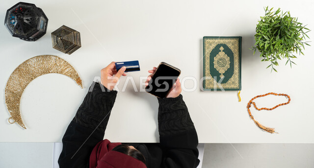 المناسبات الدينية والأعياد الإسلامية، الإستعداد لقدوم شهر رمضان المبارك، الطلب عبر المواقع الالكترونية، بورتريه علوي عامودي ليدي إمرأة عربية خليجية سعودية تستخدم الهاتف بجانبها المصحف الشريف والفوانيس والهلال، الدفع عن طريق بطاقة الصراف البنكية، خلفية بيضاء