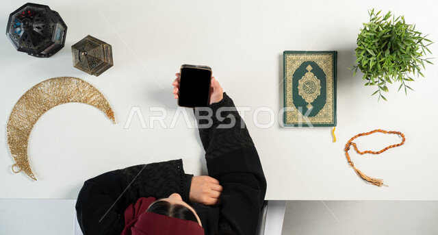 الإستعداد لقدوم شهر رمضان المبارك، المناسبات الدينية والأعياد الإسلامية، الطلب عبر المواقع الالكترونية، بورتريه علوي عامودي ليدي إمرأة عربية خليجية سعودية تستخدم الهاتف المحمول بجانبها المصحف الشريف والفوانيس والهلال، البيع والشراء عبر الانترنت، خلفية بيضاء