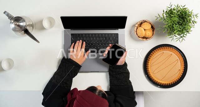 البحث عن الحلويات عن طريق المواقع الالكترونية، تشكيلة متنوعة من المأكولات الغنية بالسكريات والسعرات الحرارية، بورتريه علوي عامودي ليدي إمرأة عربية خليجية سعودية تقوم باستخدام الحاسوب والهاتف المحمول وبجانبها أنواع مختلفة من أطباق الحلو، خلفية بيضاء