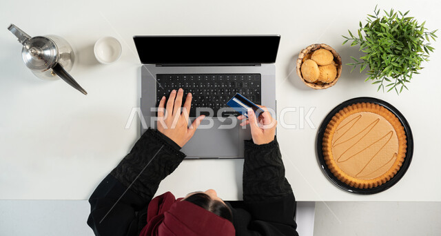 الدفع عبر بطاقة المصرف البنكية، تصفح المواقع الالكترونية للبحث عن الحلويات، تشكيلة متنوعة من المأكولات الغنية بالسكريات والسعرات الحرارية، بورتريه علوي عامودي ليدي إمرأة عربية خليجية سعودية تستخدم الحاسوب وبجانبها أنواع مختلفة من أطباق الحلو، خلفية بيضاء