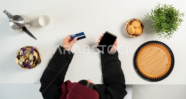 تصفح المواقع الالكترونية للبحث عن الحلويات، تشكيلة متنوعة من المأكولات الغنية بالسكريات والسعرات الحرارية، بورتريه علوي عامودي ليدي إمرأة عربية خليجية سعودية تقوم باستخدام الهاتف بجانبها أنواع مختلفة من أطباق الحلو، الدفع عبر بطاقة المصرف البنكية ، خلفية بيضاء