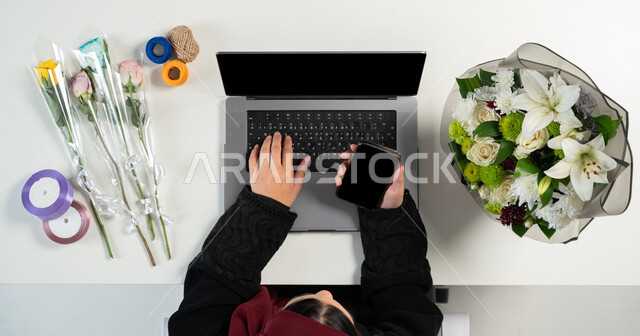 التسوق الالكتروني عبرالنت، تغليف ولف بوكيهات الزهور، بورتريه علوي عامودي ليدي إمرأة عربية خليجية سعودية تقوم باستخدام الحاسوب والهاتف، الحصول على أدوات الورود عن طريق الأونلاين، استخدام التكنولوجيا الحديثة المتطورة، عرض شاشة سوداء فارغة، خلفية بيضاء