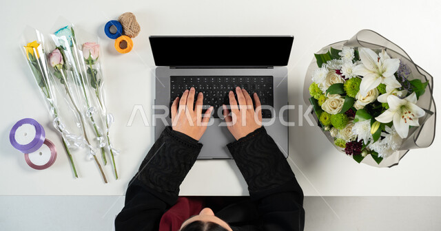 البيع والشراء عبر الانترنت، الاستفادة من التكنولوجيا الحديثة المتطورة، تغليف ولف بوكيهات الزهور، الحصول على أدوات الورود عن طريق الأونلاين، بورتريه علوي عامودي ليدي إمرأة عربية خليجية سعودية تقوم باستخدام الحاسوب المحمول، عرض شاشة سوداء فارغة، خلفية بيضاء