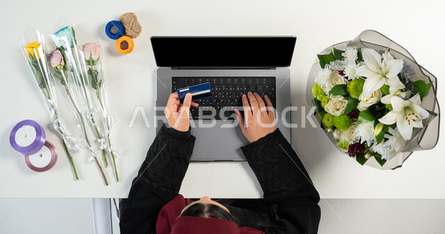 الدفع عن طريق بطاقة الصراف البنكية الأئتمانية، الحصول على أدوات الورود أونلاين، تغليف ولف بوكيهات الزهور، بورتريه علوي عامودي ليدي إمرأة عربية خليجية سعودية تقوم باستخدام الحاسوب المحمول، البيع والشراء عبر الانترنت، عرض شاشة سوداء فارغة، خلفية بيضاء