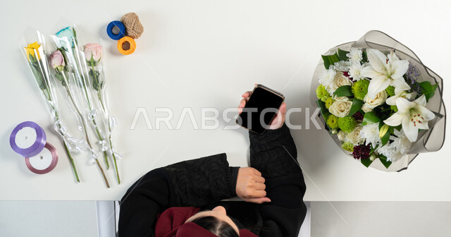 Wrapping and wrapping flower bouquets, a vertical overhead portrait of a Saudi Gulf Arab woman using a mobile phone next to her, flower supplies, buying and selling online, obtaining flower supplies online, displaying a blank black screen, white background