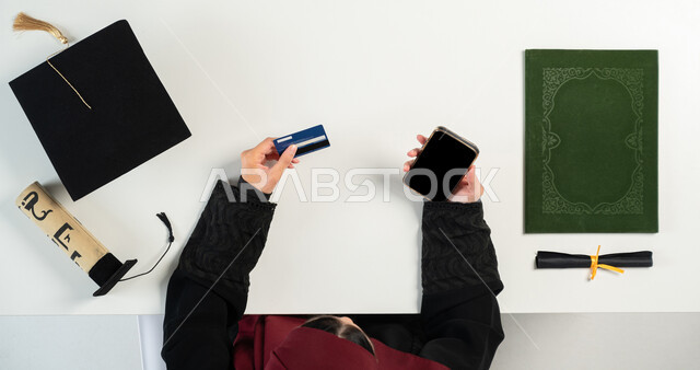 الاستفادة من التكنولوجيا الحديثة لطلب مستلزمات التخرج عن طريق الأونلاين، استخدام تطبيقات التسوق عن بعد، البيع والشراء عبر الانترنت، بورتريه علوي عامودي ليدي إمرأة عربية خليجية سعودية تستخدم الهاتف، الدفع عبر البطاقة البنكية، عرض شاشة سوداء فارغة، خلفية بيضاء