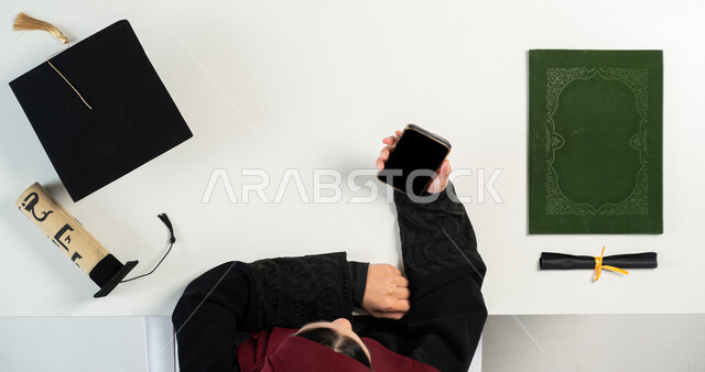 Using remote shopping applications, taking advantage of modern technology to order graduation supplies online, buying and selling online, a vertical overhead portrait of a Saudi Arabian Gulf woman using a mobile phone, displaying a blank black screen, a white background.