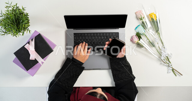 Using advanced modern technologies, buying and selling online, a vertical portrait of a lady, a Saudi Arabian Gulf woman using a computer and mobile phone, with gifts and flowers next to her, shopping through websites, displaying a blank black screen, a white background.