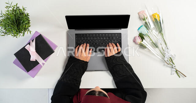 Taking advantage of advanced modern technology, buying and selling online, a vertical overhead portrait of a Saudi Gulf Arab woman using a laptop with gifts and flowers next to her, shopping through websites, displaying a blank black screen, white background