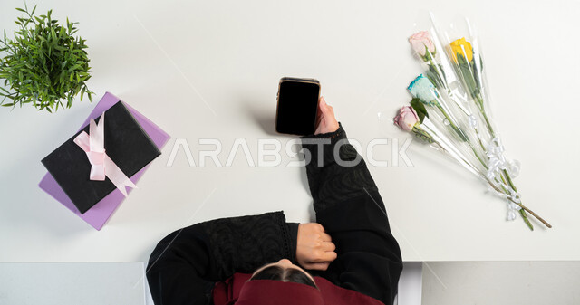 البيع والشراء عبر الانترنت، مفهوم التسوق الالكتروني، بورتريه علوي عامودي ليدي إمرأة عربية خليجية سعودية تستخدم الهاتف المحمول وبجانبها مجموعة من الهدايا و الأزهار، استخدام التقنيات الحديثة المتطورة، عرض شاشة سوداء فارغة، خلفية بيضاء