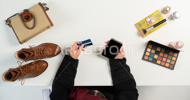 Getting personal items and cosmetics online, e-shopping concept, vertical top portrait of a Saudi Arabian Gulf woman paying bills via mobile phone using bank card, integrating modern technologies into daily life, white background