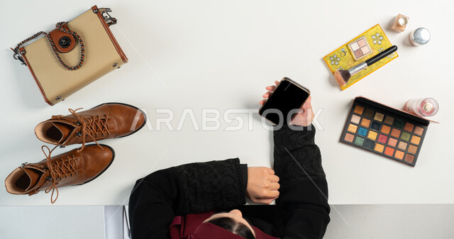 مفهوم التسوق الإلكتروني، الحصول على الأغراض الشخصية ومستحضرات التجميل عن طريق الأونلاين، بورتريه علوي عامودي ليدي إمرأة عربية خليجية سعودية تقوم بتصفح العروضات على المواقع الالكترونية عبر الهاتف المحمول، دمج التقنيات الحديثة في الحياة اليومية، خلفية بيضاء