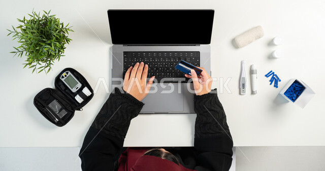 Using a bank card to obtain medical devices and supplies online, the concept of an electronic clinic, medical consultations via websites, vertical top portrait of a Saudi Arabian Gulf woman using a laptop, displaying a blank black screen, white background