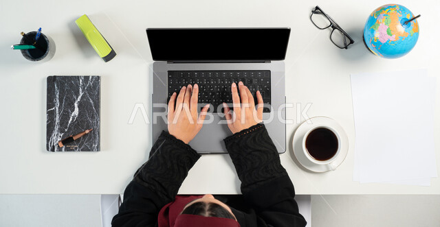البيع والشراء عبر الانترنت، بورتريه علوي عامودي لإمرأة عربية خليجية سعودية تستخدم جهاز الحاسوب المحمول وبجانبها مجموعة من القرطاسية، التسوق من خلال المواقع الإلكترونية، الاستفادة من التكنولوجيا للحصول على الكتب والدفاتر أونلاين، عرض شاشة سوداء فارغة، خلفية بيضاء