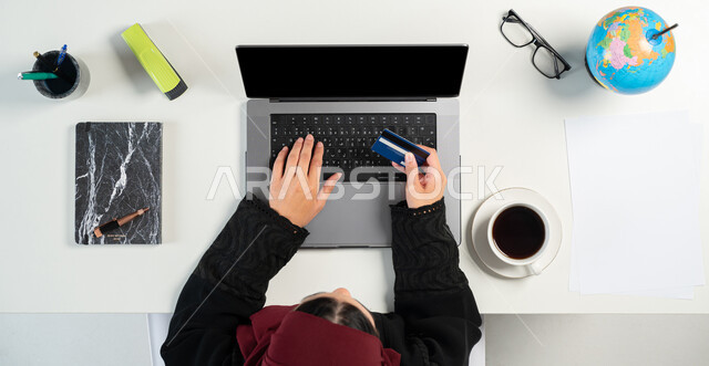 الحصول على الكتب والدفاتر أونلاين، عرض شاشة سوداء فارغة، استخدام البطاقة النبكية للبيع والشراء عبر الانترنت، التسوق من خلال المواقع الإلكترونية، بورتريه علوي عامودي لإمرأة عربية خليجية سعودية تستخدم الحاسب المحمول بجانبها مجموعة من القرطاسية، خلفية بيضاء