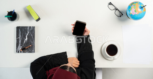 البيع والشراء عبر الانترنت، التسوق من خلال المواقع الإلكترونية، بورتريه علوي عامودي لإمرأة عربية خليجية سعودية تستخدم الهاتف المحمول بجانبها مجموعة من القرطاسية، الاستفادة من التكنولوجيا للحصول على الكتب والدفاتر أونلاين، عرض شاشة سوداء فارغة، خلفية بيضاء