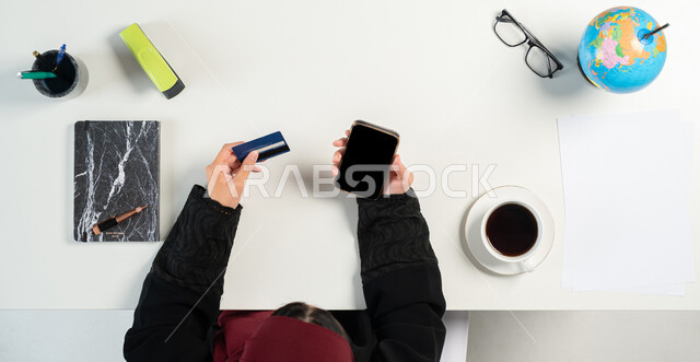 استخدام البطاقة النبكية للبيع والشراء عبر الانترنت، التسوق من خلال المواقع الإلكترونية، بورتريه علوي عامودي لإمرأة عربية خليجية سعودية تستخدم الهاتف المحمول بجانبها مجموعة من القرطاسية، الحصول على الكتب والدفاتر أونلاين، عرض شاشة سوداء فارغة، خلفية بيضاء