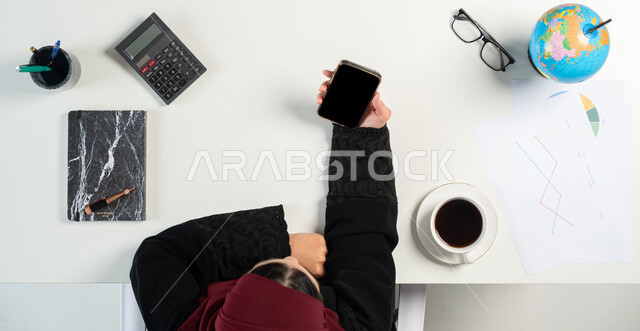 إستخدام الآلة الحاسبة لإجراء العمليات الحسابية ودراسة المشاريع، العمل في قسم التحليلات والإحصائيات البيانية، بورتريه علوي عامودي لإمرأة عربية خليجية سعودية تعمل على إدارة الحسابات بإستخدام الهاتف المحمول، مهن ووظائف نسائية مكتبية، خلفية بيضاء
