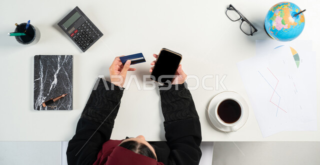 إدارة الحسابات بإستخدام الهاتف المحمول، إستخدام الآلة الحاسبة لإجراء العمليات الحسابية ودراسة المشاريع، العمل في قسم التحليلات والإحصائيات البيانية، بورتريه علوي عامودي لإمرأة عربية خليجية سعودية تحمل بيدها البطاقة الائتمانية، مهن ووظائف نسائية مكتبية، خلفية بيضاء
