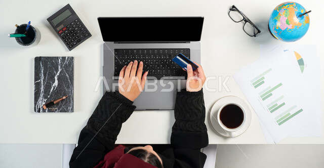 Financial investment by bank card, following the economic growth chart through technical devices, monitoring the economy and financial information, vertical top portrait of a Saudi Arabian Gulf woman studying and analyzing statistics and charts on a laptop, white background