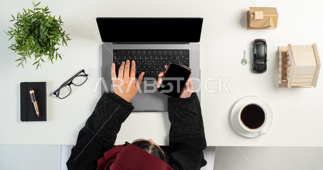 العمل في المجال العقاري عبر الحاسب المحمول، تصفح مواقع العقارات الالكترونية, شراء عقار جديد عبر الاونلاين, بورتريه علوي عامودي لإمرأة عربية خليجية سعودية تحمل جوال بشاشة سوداء فارغة، مجسمات صغيرة للمنازل، امتلاك سكن جديد، مفهوم الاسكان والتمليك، خلفية بيضاء