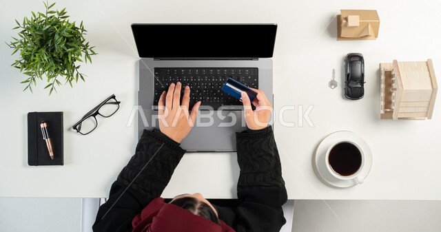 استخدام البطاقة البنكية لشراء عقار جديد عبر الاونلاين, تصفح مواقع العقارات الالكترونية, بورتريه علوي عامودي لإمرأة عربية خليجية سعودية تعمل في المجال العقاري عبر الحاسب المحمول، مجسمات صغيرة للمنازل، امتلاك سكن جديد، مفهوم الاسكان والتمليك، خلفية بيضاء