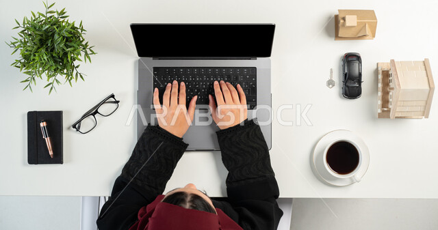 تصفح مواقع العقارات الالكترونية, استخدام الوسائل التقنية لشراء عقار جديد عبر الاونلاين, بورتريه علوي عامودي لإمرأة عربية خليجية سعودية تعمل في المجال العقاري عبر الحاسب المحمول، مجسمات صغيرة للمنازل، امتلاك سكن جديد، مفهوم الاسكان والتمليك، خلفية بيضاء