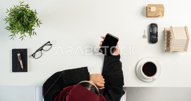 تصفح مواقع العقارات الالكترونية, استخدام الوسائل التقنية لشراء عقار جديد عبر الاونلاين, بورتريه علوي عامودي لإمرأة عربية خليجية سعودية تعمل في المجال العقاري عبر الهاتف المحمول، مجسمات صغيرة للمنازل، امتلاك سكن جديد، مفهوم الاسكان والتمليك، خلفية بيضاء