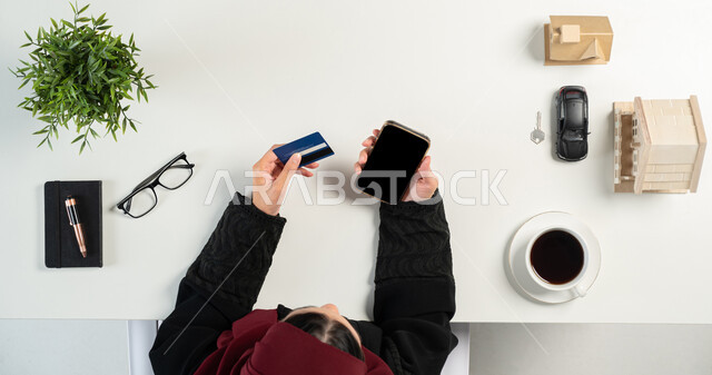 مفهوم الاسكان والتمليك، استخدام البطاقة البنكية لشراء عقار جديد عبر الاونلاين, تصفح مواقع العقارات الالكترونية, بورتريه علوي عامودي لإمرأة عربية خليجية سعودية تعمل في المجال العقاري عبر الهاتف المحمول، مجسمات صغيرة للمنازل، امتلاك سكن جديد، خلفية بيضاء