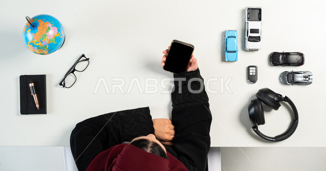 Shopping for music listening devices online, vertical top portrait of a Saudi Arabian Gulf woman using a mobile phone, online buying and selling, getting modern and advanced technologies online, displaying a blank black screen on a mobile phone, white background