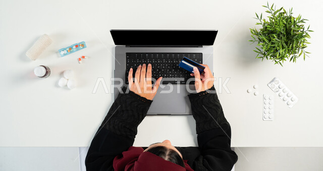 التسوق عبر المواقع الإلكترونية، بورتريه علوي عامودي ليدي امرأة عربية خليجية سعودية تقوم باستخدام الحاسوب المحمول للدفع عن طريق بطاقة الصراف البنكية, الحصول على مجموعة من الأدوية عبر الانترنت، شراء أنواع من الدواء أونلاين، عرض شاشة سوداء فارغة، خلفية بيضاء