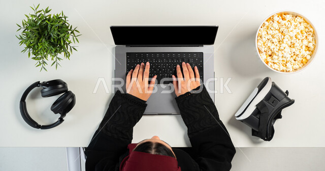 التسلية والاستمتاع بالاستراحة، البيع والشراء أونلاين، نظارة الواقع الافتراضي مع سماعة الرأس وطبق من الفشار على طاولة بيضاء، بورتريه مقرب ليدي إمرأة عربية خليجية سعودية تستخدم جهاز الحاسوب المحمول وبجانبها طبق من الفوشار، عرض شاشة سوداء فارغة، خلفية بيضاء