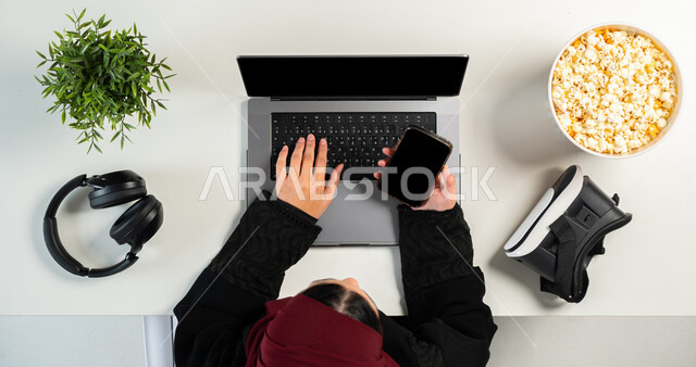 Virtual reality glasses with a headset and a bowl of popcorn on a white table, the concept of entertainment with technology and modern technology, payment by bank card for purchases via mobile phone and laptop, close-up portrait of a Saudi Gulf Arab woman using a mobile phone, displaying a blank black screen, white background