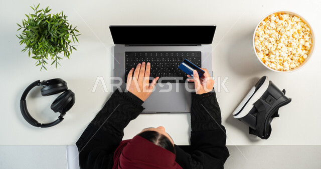 مفهوم الترفيه والتسلية بالتقنية والتكنولوجيا الحديثة، نظارة الواقع الافتراضي مع سماعة الرأس وطبق من الفشار على طاولة بيضاء، الدفع بالبطاقة البنكية للشراء عبر الحاسوب المحمول، بورتريه علوي عامودي ليدي إمرأة عربية خليجية سعودية تستخدم اللابتوب، عرض شاشة سوداء فارغة، خلفية بيضاء