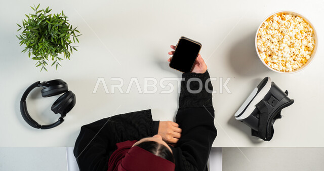 نظارة الواقع الافتراضي مع سماعة الرأس وطبق من الفشار على طاولة  بيضاء، مفهوم الترفيه والتسلية بالتقنية والتكنولوجيا الحديثة، الدفع بالبطاقة البنكية للشراء عبر الهاتف المحمول، بورتريه علوي عامودي ليدي إمرأة عربية خليجية سعودية تستخدم الجوال، عرض شاشة سوداء فارغة، خلفية بيضاء