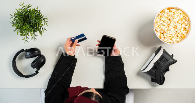 قضاء الوقت عبر مشاهدة فيلم من خلال نظارات الواقع الإفتراضي ، بورتريه علوي عامودي ليدي إمرأة عربية خليجية سعودية تستخدم الجوال للدفع عن طريق البطاقة البنكية وبجانبها طبق من الفوشار، التسلية والاستمتاع بالاستراحة، البيع والشراء أونلاين، عرض شاشة سوداء فارغة، خلفية بيضاء