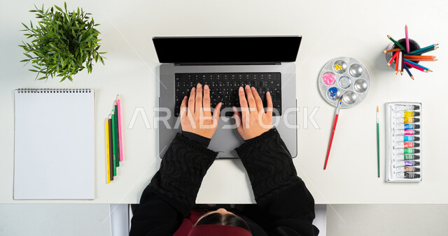 التعلم والتدريب من خلال المواقع الإلكترونية، بورتريه ليدي إمرأة عربية خليجية سعودية تقوم باستخدام اللابتوب وبجانبها أقلام الرسم والتلوين، البيع والشراء عبر الانترنت، الحصول على الألوان المائية والخشبية ومستلزمات الرسم أونلاين، عرض شاشة سوداء فارغة، خلفية بيضاء