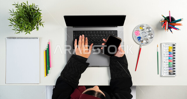 البيع والشراء عبر الانترنت، التعلم والتدريب من خلال المواقع الإلكترونية، بورتريه ليدي إمرأة عربية خليجية سعودية تقوم باستخدام الجوال وجهاز الحاسوب المحمول وبجانبها أدوات الرسم، الحصول على مستلزمات الرسم أونلاين، عرض شاشة سوداء فارغة، خلفية بيضاء