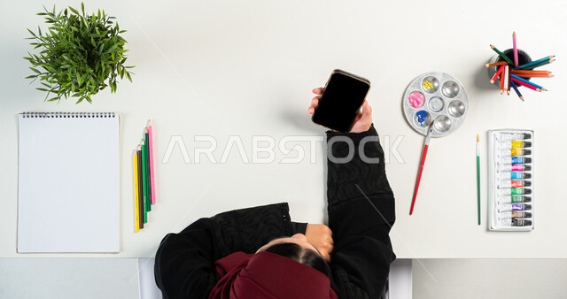 التعلم والتدريب من خلال المواقع الإلكترونية، بورتريه ليدي إمرأة عربية خليجية سعودية تقوم باستخدام الهاتف الذكي وبجانبها ألوان وأدوات الرسم، البيع والشراء عبر الانترنت، الحصول على مستلزمات وأغراض الرسم أونلاين، عرض شاشة سوداء فارغة، خلفية بيضاء